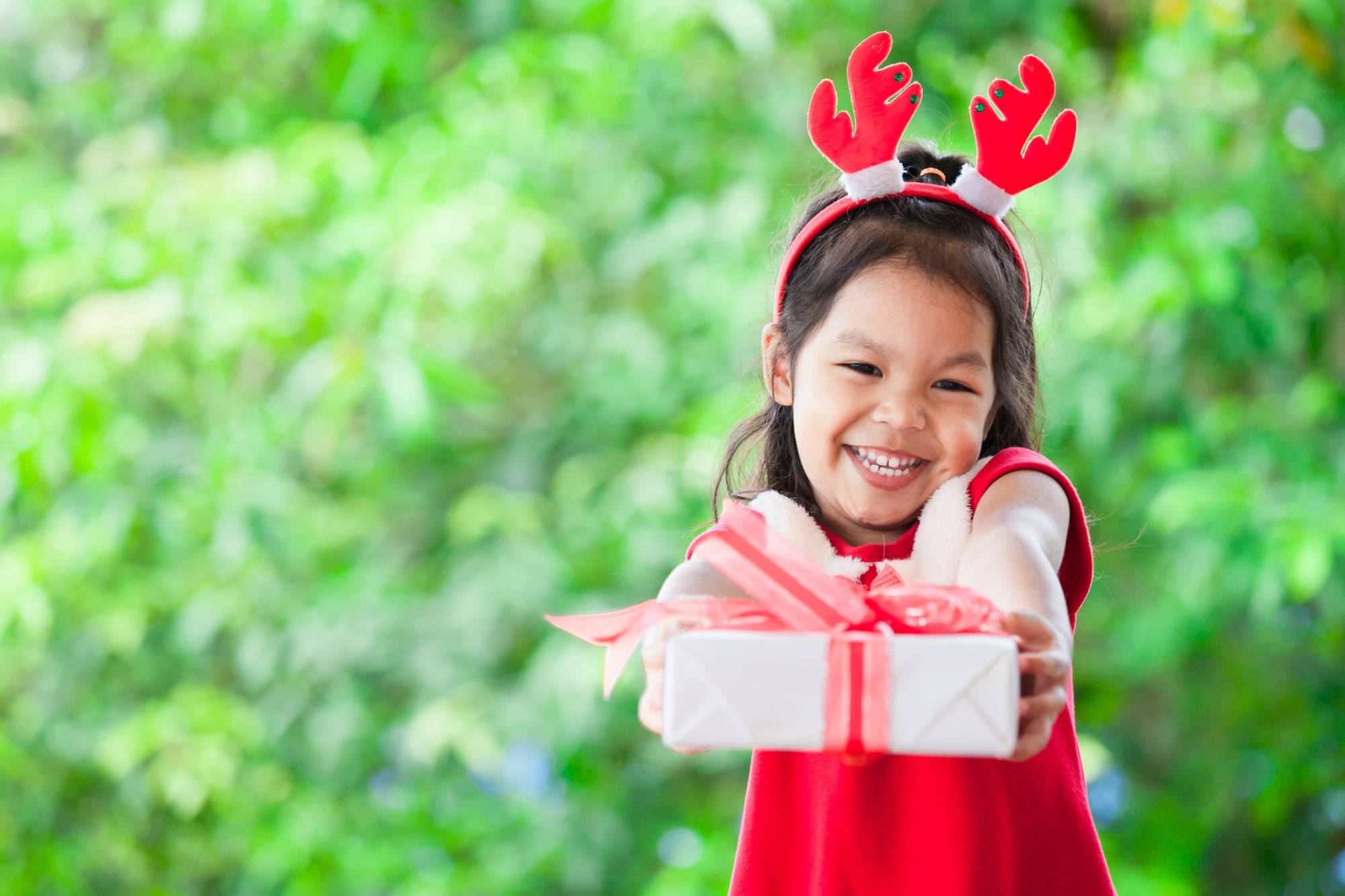Cute asian child girl holding and giving Christmas gift on green nature background Cute asian child girl holding and giving Christmas gift on green nature background